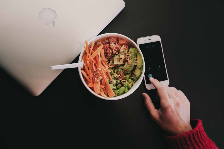 A bowl of fresh salad with avocado, carrots, edamame, and quinoa on a desk beside a smartphone and laptop.