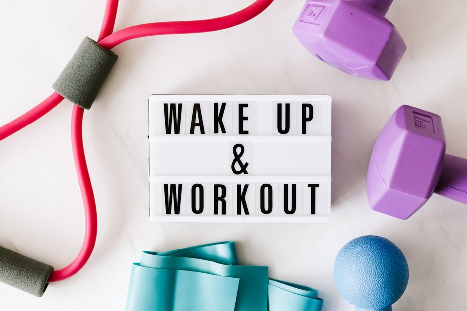 Top view closeup composition of wake up and workout inscription on light box surface among expanders and dumbbells and massage ball on white background