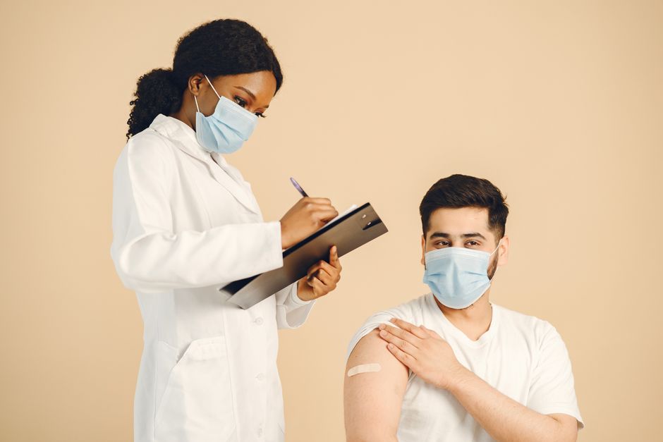 Healthcare professional wearing mask administering a vaccine to a patient in a beige studio setting.