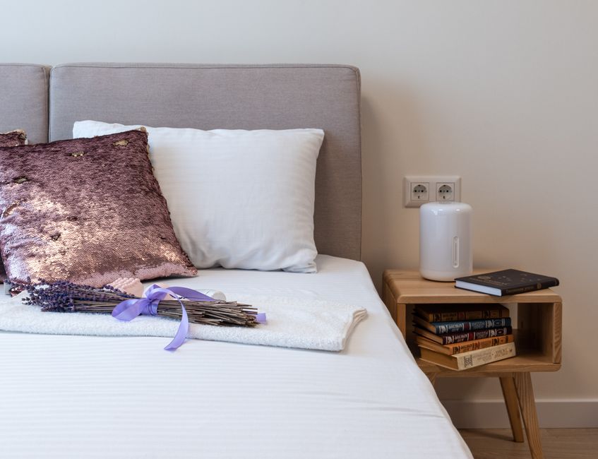 Bunch of fresh aromatic lavender placed on comfortable bed with decorative pillows near wooden night table with stack of books