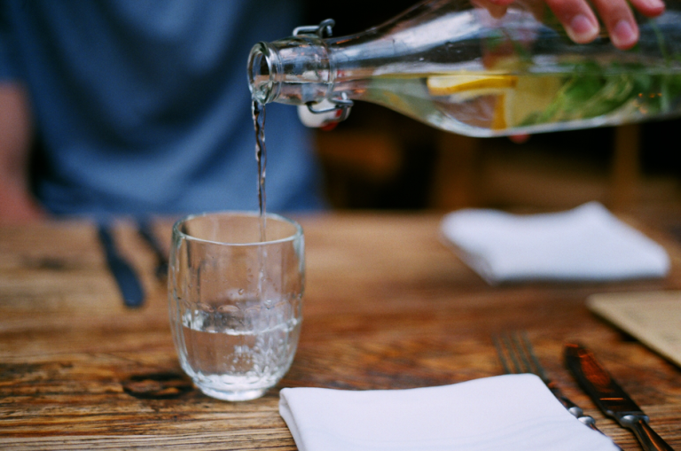A hand pours lemon infused water from a glass bottle into a drinking glass on a rustic wooden table.